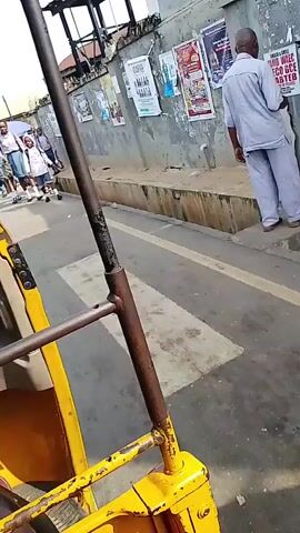 The video is filmed from the perspective of someone inside a yellow, three-wheeled vehicle, likely a tuk-tuk or auto-rickshaw. The vehicle is moving along a paved road in what appears to be a town or city. On the right side of the road, there is a sidewalk with a concrete wall behind it. The wall is covered with various posters.

Several people are walking on the sidewalk. In the foreground on the sidewalk, there are a group of young people, likely students, wearing school uniforms. Some of them are carrying backpacks. They are walking in the same direction as the vehicle. Further down the sidewalk, there are other adults and children. One adult is wearing a blue and white checkered shirt, another is wearing a dark colored top and a red skirt, and a man in a blue and white patterned shirt stands near a car. There are also market stalls with umbrellas visible in the background.

The overall environment suggests a busy street scene in a developing country. The road is paved but appears worn. The buildings lining the street are simple structures. The weather appears to be daytime with sunlight casting shadows. The video seems to capture a moment of daily life, possibly the commute of students to or from school.