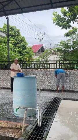 An exterior shot of two men by a wall with faucets and a large blue water barrel. The man on the left, wearing a light brown collared shirt and a white cap, stands facing the camera, resting his left elbow on the barrel. The man on the right, wearing a blue t-shirt and grey pants, is bent over at the faucets, washing his hands. The area is covered by a roof and paved with patterned tiles, with a drain in the foreground. Trees and houses are visible in the background under a cloudy sky.
