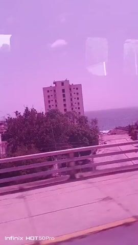 A pink-tinted video captures a moving view from inside a vehicle, likely a bus, as it travels along a road. The road is visible in the foreground, with a guardrail running along its edge. Beyond the road, the landscape consists of trees and buildings, including a tall, light-colored apartment building with visible windows and balconies. The ocean or a large body of water can be seen in the background, under a sky with scattered clouds. The camera appears to be looking out a window, with reflections visible on the glass. The video appears to be a short, continuous shot with no notable action or interaction occurring within the frame. The overall impression is a snapshot of urban and coastal scenery during daytime. The timestamp indicates the video is from January 1970, which is likely a default setting for the recording device. The watermark at the bottom of the screen reads "Infinix HOT 50 Pro".