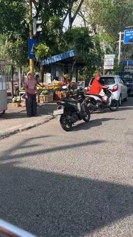 The video shows a street scene with several people and vehicles. In the foreground, a black motorcycle is parked on the side of the road. Behind it, a white car is also parked. On the sidewalk, a woman in a red hijab is sitting on a scooter. Further back, another woman in a pink hijab is standing near a fruit stand. The fruit stand is laden with various fruits, including watermelons. The environment appears to be an urban or semi-urban area, with trees and buildings visible in the background. The lighting suggests it is daytime, possibly late afternoon given the long shadows cast on the road.