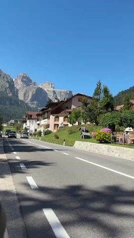 A daytime exterior scene captures a winding road with a white line marking its lanes, leading through a village nestled at the base of a mountain range. On the right side of the road, a gently sloping green hill is dotted with trees, flowering bushes, and the facades of several houses. Some houses are pink and white, with balconies and roofs visible. On the left side of the road, there are some parked cars and more houses lining the street. A few cars are driving on the road, appearing to move away from the viewer. The sky is a clear, bright blue. The mountains in the background are rugged and appear to be covered in some vegetation at their base, with rocky, bare peaks reaching towards the sky.
