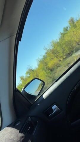 The video shows the interior of a car with a view out of the side window. The sky is clear and blue. On the right side of the frame, a side mirror is visible, reflecting a road and trees. The trees appear to be green and yellow, possibly indicating a change of season. The car's interior, including the door panel and air vent, is also visible. The perspective suggests the car is in motion.