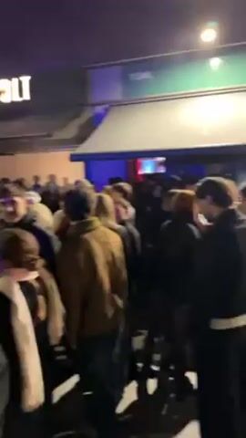 The video shows a large crowd of people gathered on a city street at night. The crowd appears to be densely packed, with individuals standing close together. Most people are wearing dark clothing. Some individuals are conversing in small groups, while others are looking around. Streetlights illuminate the scene, and there's a building with a "WOLT" sign visible. In the background, there is a dark building and a light with a purple hue.