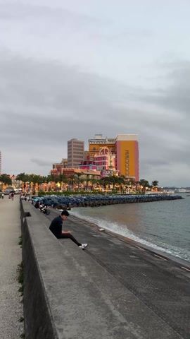 The video shows a medium shot of a waterfront scene. A person wearing dark clothing sits on a concrete ledge on the left side of the frame, looking at their phone. In the background, a colorful, multi-story building with a mix of architectural styles stands out. The building is illuminated with warm lights, adding a vibrant contrast to the overcast sky. Along the shoreline, there are palm trees decorated with lights. Other people are visible in the distance, walking along the waterfront. The sea gently laps against a row of concrete breakwaters. The overall atmosphere is calm and serene.