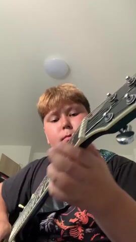 A close-up shot shows a young boy playing an electric guitar indoors. He has blond and brown hair styled in a quiff and is wearing a black t-shirt with a graphic design featuring skeletons. The boy's face is focused as he strums the guitar. The background reveals a white ceiling with a circular light fixture, and a closet door with a poster on it. The lighting suggests an indoor environment, likely a bedroom, with natural light entering from an unseen source.