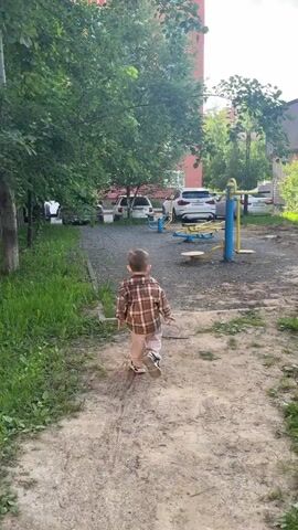 The video shows a young child from behind, walking on a dirt path in an outdoor setting. The child is wearing a brown and white/beige plaid long-sleeved shirt, light-colored trousers, and sneakers. They walk across a gravel area towards blue and yellow outdoor exercise equipment, eventually stopping near one of the machines.

The scene appears to be a residential courtyard or park, possibly in Odintsovo, Russia, as indicated by the context. Lush green trees line the path, and several cars (white, grey, silver SUVs and sedans) are parked in the background beyond the exercise area. A multi-story building, possibly an apartment complex with red-toned walls, is partially visible through the trees. The ground is a mixture of dirt, gravel, and grass. The weather is bright and sunny, indicating daytime. No visible text is present in the video.