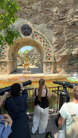 The video shows a group of people standing on a paved walkway in front of a water attraction. In the background, there is a large, ancient-looking stone archway with a fierce face carved into it. Beyond the archway, a golden statue stands on a pedestal. The water in the attraction appears to be murky and orange. Two cannons are positioned on the walkway, and several people are holding them. In the foreground, a woman in a black tank top and white pants is looking at the water. To her left, a woman in a dark blue hijab and another woman in a purple shirt are also looking towards the attraction. To the right of the woman in white pants, a person wearing a light green t-shirt and a backpack is also facing the attraction. The scene is outdoors, with a rock face on the right and trees with green leaves visible on the left. The sky is bright and appears to be sunny.