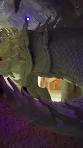 The video shows a close-up view of a large, grey, dragon-like sculpture inside what appears to be a cave or grotto. The sculpture is highly detailed, with scales covering its body and intricate facial features, including fangs and elaborate headdresses. In the background, two individuals are visible. One, wearing a red shirt and black pants, is partially obscured by the sculpture. The other, wearing a black t-shirt, walks past the sculpture. The environment is dimly lit, with purple and blue lights illuminating parts of the cave and the sculpture, creating a mystical atmosphere. The camera slowly pans and zooms, offering different perspectives of the sculpture and the surrounding environment.