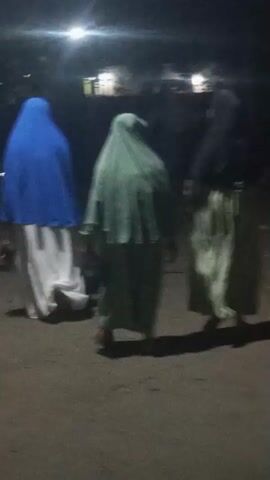 A group of individuals, appearing to be women wearing hijabs and long dresses, walk away from the camera on a dark, unpaved surface. The lighting suggests it is nighttime or dusk. The individual on the far left wears a blue hijab and a white dress. The person next to them has a light green hijab and dress. The individual on the far right wears a dark hijab and a light green dress. Their faces are not visible. The ground appears to be dirt or gravel. In the background, there are indistinct shapes of what could be buildings or trees, illuminated by a distant light source. The overall scene is dark and somewhat blurry.