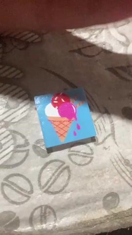 A close-up, overhead shot shows a small, square sticker with an image of an ice cream cone on a blue background. The ice cream cone has two scoops of ice cream, one red and one pink, and is dripping. The sticker is placed on a textured, light-colored surface that appears to be fabric or a similar material with a repeating pattern of circles with lines through them. The lighting creates a shadow across the upper portion of the image, obscuring some of the background detail. The sticker appears to be a few inches in size.
