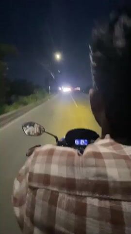 A person wearing a brown and white plaid shirt drives a motorcycle at night, filmed from a first-person perspective behind the driver. The motorcycle's digital dashboard is visible, as is the right rearview mirror showing a blurred reflection of the driver. The motorcycle travels on an asphalt road with a yellow solid center line. Oncoming vehicle headlights create a bright glare in the distance, illuminating the road ahead with a yellowish glow. A streetlight is visible high on the left. On the left side of the road, a white barrier or railing is passed. The distinct sound of a vehicle honking is audible at the beginning of the clip.
