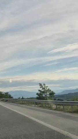 The video shows a view from a moving vehicle on a paved road. A metal guardrail runs along the side of the road. In the distance, there are rolling hills and mountains under a sky with scattered clouds. Trees are visible along the side of the road. The video is shot in landscape orientation.