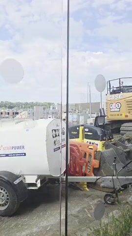 The video shows a construction site. In the foreground, there's a white tank with hazard symbols, a red barrier, and construction equipment including a yellow compactor and a large excavator with a bucket. In the background, there are unfinished buildings, construction materials like pipes and cones, and a white spherical object suspended in the air. The sky is cloudy. A "No Smoking" sign is visible on a window. The camera pans across the scene.