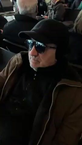 The video shows a man in an airport waiting area. He is wearing sunglasses, a black cap with ear flaps, a brown jacket with a dark fleece collar, and a black zip-up shirt. He is sitting down and holding a wooden cane in his right hand. Other people are visible in the background, sitting and walking. There is a window behind the man, and an airline logo is visible on the wall.