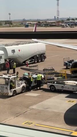 The video captures a scene at an airport tarmac on a bright, sunny day. In the foreground, a small white tow truck with the number "32" visible on its side is positioned near a medical stretcher. Several individuals, some wearing high-visibility vests, are actively engaged in transferring equipment onto a luggage cart. The stretcher appears to be part of an operation involving medical assistance or transport of a patient.

In the background, a large white tanker truck labeled "JET A-1 NO.6" is parked. Beyond the immediate activity, the vast expanse of the airport runway and surrounding infrastructure is visible, including distant buildings and a control tower. The presence of an airplane wing in the upper right corner of the frame further emphasizes the airport setting. The overall impression is one of organized airport operations, possibly with an emphasis on passenger safety or medical support. The clear sky and bright sunlight suggest a warm, pleasant weather condition.
