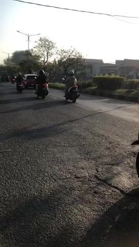 The video shows a street scene with several motorcycles and riders. The camera is positioned behind a motorcycle with two people on it. The rider in front is wearing a helmet, and the passenger behind is also wearing a helmet. They are traveling on a paved road with other motorcycles in front of them, also carrying passengers. The sun is low in the sky, casting long shadows. There are trees and buildings visible in the background. The overall impression is of a commute or a journey on a sunny day.