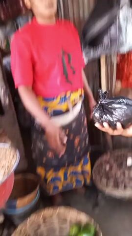 A person in a red t-shirt and a patterned skirt stands inside a market stall. The stall has various items for sale including beans, nuts, tomatoes and onions. Another person in a white t-shirt and blue shorts approaches the stall, pays with a phone and is handed a black plastic bag.
