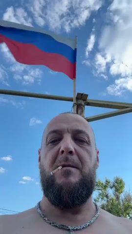 A low-angle, close-up, selfie-style video features a bald, bearded man with a cigarette in his mouth. Behind him, a Russian flag flutters on a flagpole against a bright blue sky with scattered clouds. The man is shirtless and wears a chain necklace. The flagpole appears to be mounted on a rusty, metal structure, possibly part of a fence or scaffolding. The overall scene suggests an outdoor setting on a sunny day.