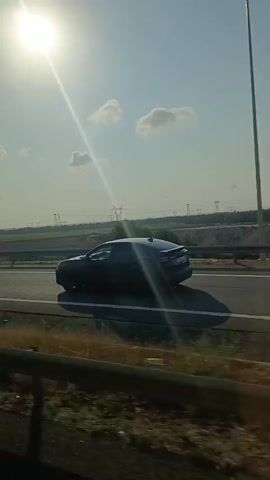 The video shows a view from a moving vehicle, looking out at a highway under a bright, sunny sky with scattered clouds. The sun is prominently visible, casting a strong glare. The highway has multiple lanes and a concrete barrier. To the side of the highway, there are dry grassy areas and scattered trees. In the distance, there are agricultural fields and power lines. A black car is seen driving on the highway in the same direction as the viewer. The perspective is consistent throughout the video, suggesting it was filmed from a single vantage point within the vehicle.