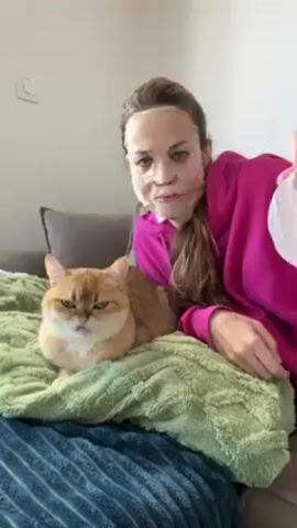 A woman with long brown hair and a pink sweatshirt lies on a couch with a green blanket, next to a ginger cat. She has a transparent face mask on and is holding a white paper face mask up to the cat's face. The paper mask has cutouts for the eyes and a mouth. The cat looks grumpy. There is a grey pillow behind the woman, and a wall with a light switch in the background.
