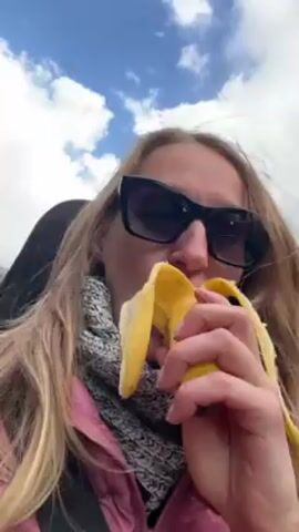 A woman with long blonde hair, sunglasses, a pink jacket, and a gray scarf is eating a banana while riding in a convertible. The car is traveling on a mountain road with bare trees on the hillside. The sky is blue with white clouds. Music can be heard in the background.