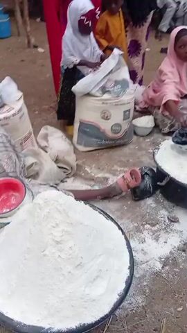 A high-angle outdoor video shows several people, including children, involved in what appears to be a food distribution or preparation event. The ground is dirt, partially covered with spilled white powder, likely flour. In the foreground, a large black basin is filled with white powder, with several small bowls stacked on top. Another basin filled with white powder is visible further back, with a blue scoop resting on the powder. A person in a pink headscarf and patterned dress is seated on the ground, scooping white powder from a basin. To the left, a child in a white headscarf and patterned dress is kneeling next to a large white sack, appearing to handle its contents. Another child in an orange top is also near the sack. The background shows more people and what looks like an outdoor gathering.