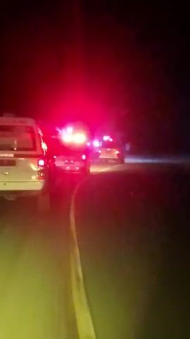 The video shows a nighttime scene on a road. In the foreground, the rear of a white ambulance with flashing red lights is visible. To the right of the ambulance, another vehicle with flashing red lights is present, and further down the road, a third vehicle is also visible with its lights on. The road appears to have a yellow line marking. The background is dark, suggesting it is night, with some lights visible in the distance on the right side of the road. The overall impression is one of an emergency response or traffic incident occurring on a road at night.