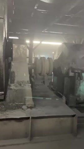 The video shows an industrial setting, possibly a factory or processing plant. The environment appears dusty and industrial.  There are large pieces of machinery, including what looks like rollers or presses, gears, and pipes.  Some machinery is covered in dust and appears to be in operation or recently used, with some dark liquid or debris collected in buckets or chutes.  The lighting is industrial, with some bright lights in the background, possibly indicating a working area.  There are no people visible in the video. The overall impression is one of an active, albeit somewhat grimy, industrial workspace.