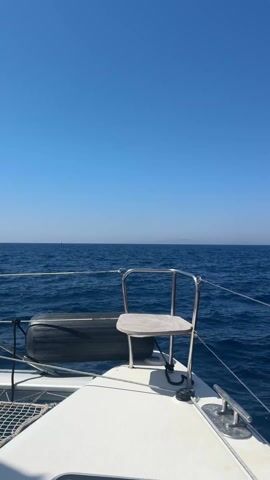 The video, captured from the bow of a boat, opens with a view of a vast, calm blue sea under a clear sky. A distant white sailboat can be seen on the horizon. As the camera pans right, a dramatic volcanic coastline comes into view. The cliffs display striking shades of red and dark brown rock, characteristic of the region.

The boat approaches a small port area nestled at the base of these cliffs. White buildings with red roofs are visible, along with a winding road where cars are parked. Further up the steep hillside, numerous white, traditional Cycladic-style houses are built into the cliff face, appearing to be part of a larger town or village. Several boats, including sailboats and yachts, are docked in the port. The scene depicts a sunny day with clear weather, suggesting a Mediterranean setting, likely in the Aegean Sea.