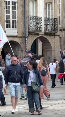 A group of people are walking through a town square. A police officer in uniform is walking through the crowd. A man is holding a flag. There is a sign that reads "Praza do Toural." The buildings in the background are made of stone and have balconies. The weather appears to be overcast.