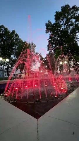This is a vertical video of a fountain at dusk. The water is illuminated by red lights, creating a vibrant effect. The fountain is surrounded by trees, and in the background, there is a building and streetlights. The sky is a gradient of blue and orange, indicating the transition from day to night. The water is spraying in various patterns, some high and thin, others wider and more dispersed. The overall impression is one of a peaceful and visually appealing outdoor scene.