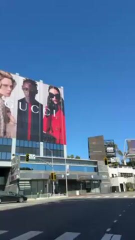 The video shows a street scene in what appears to be a commercial district. The camera pans upwards, focusing on a billboard featuring the text "wave check" and an image of a person's face. To the left, there's a large Gucci advertisement on the side of a building. Traffic lights are visible at the intersection, and a black car is parked on the side of the road. The sky is clear and blue.
