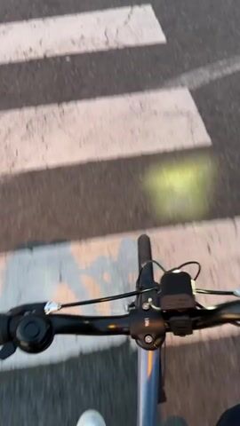 The video shows a first-person view of someone riding a bicycle across a pedestrian crosswalk. The camera is positioned at the handlebars of the bicycle. The rider is wearing white shoes and black pants. The bicycle has a light that illuminates the road. The camera then pans to the right to reveal a road, a hedge, and a view of mountains against a pink and orange sunset sky. There is a circular traffic mirror beside the road.