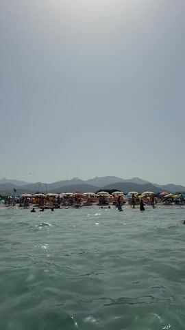 The video shows a crowded beach with many people relaxing under umbrellas and swimming in the ocean. The water is clear and blue, and the sky is bright and sunny. In the background, there are mountains. The video captures the atmosphere of a typical summer day at the beach.