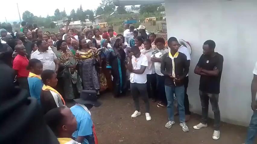 Here is a concise description of the scene:

A crowd of people has gathered outdoors near a light-colored wall. The group is diverse in age and attire. Several people stand with arms crossed, and some are wearing uniforms with neckerchiefs. In the background, there are trees and buildings, suggesting a rural or suburban setting. The group seems to be focused on something happening towards the left of the frame, as indicated by their line of sight and body language. There is a sense of anticipation, and one person is possibly speaking or leading the group, based on the open mouths visible among some members of the crowd.
