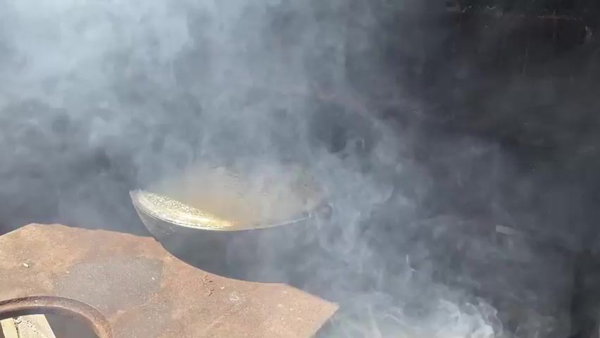 A cast iron pot with a glass lid is steaming on a metal surface. The pot is situated within a brick structure, possibly an oven or fireplace. Thick white smoke billows around the pot, obscuring parts of the background. The lighting suggests it is daytime, with sunlight casting reflections on the pot's lid. The surface the pot rests on appears weathered and possibly made of cast iron as well, with a circular opening visible in the lower left corner. The brick structure is dark and appears to be made of rough, unplastered bricks.