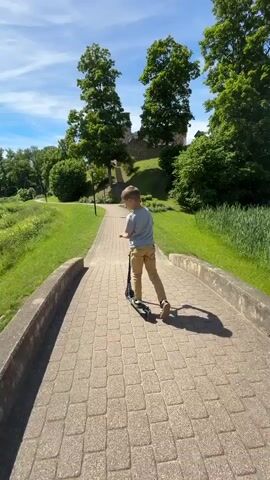 A young boy rides a scooter down a paved path in a park. The path is made of cobblestones and is lined with a low stone wall on either side. The boy is wearing a light blue t-shirt and khaki pants. He is facing away from the camera. To the left of the path is a grassy slope leading down to a body of water with a fountain. To the right of the path, there is a grassy hill with trees and the ruins of a stone structure at the top. The sky is blue with scattered white clouds, and the sun is shining. The video is shot from a low angle, looking forward.