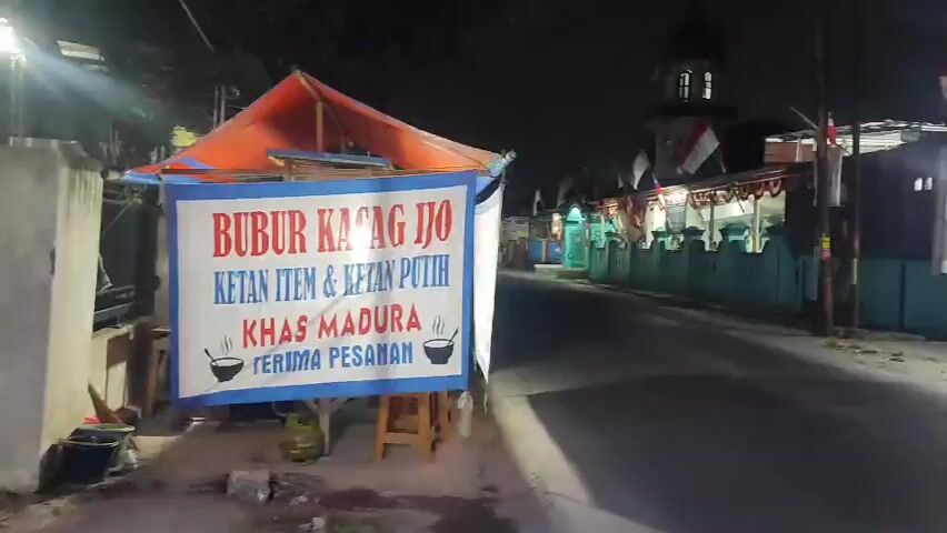 The video shows a street scene at night. On the left, a food stall displays a banner advertising "BUBUR KACANG IJO KETAN ITEM & KETAN PUTIH KHAS MADURA TERIMA PESANAN" which translates to "Green Bean Porridge Black Sticky Rice & White Sticky Rice MADURESE SPECIAL TAKEOUT ACCEPTED". The stall has an orange tarp roof and is supported by wooden legs. A gas tank is visible underneath. On the right side of the street, there are several shops and stalls, some of which are illuminated. One stall has a banner with images of apples and the text "BERAS BASMATI APPLE", suggesting it sells basmati rice. There are stacks of bottled water and other products displayed. In the background, a building with a dome, possibly a mosque, is visible, adorned with Indonesian flags. The street is paved and appears to be a public thoroughfare. The overall impression is of a roadside food and goods vendor area operating at night.