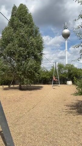 A young person in red swim trunks is swinging on a zipline in an outdoor area with a large tree and a water tower in the background. The zipline equipment includes metal poles and cables. The ground is covered in wood chips. The person is swinging back and forth, their body extending out from the zipline as they swing. The sky is partly cloudy.