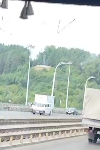 The video pans across a bridge with traffic. In the foreground, there are train tracks. On the bridge, a white van, a white box truck, a blue articulated bus, a dark SUV, and a white car are visible. In the background, there is a densely wooded hill with a statue on top, illuminated by streetlights. The sky is overcast.