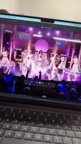 A person is recording the screen of a laptop showing a pop singer performing with dancers on stage with pink and purple lighting and pyrotechnics. The screen then cuts to a fireworks show over a city. The person holding the laptop is indoors. A man's head is briefly visible in the background.
