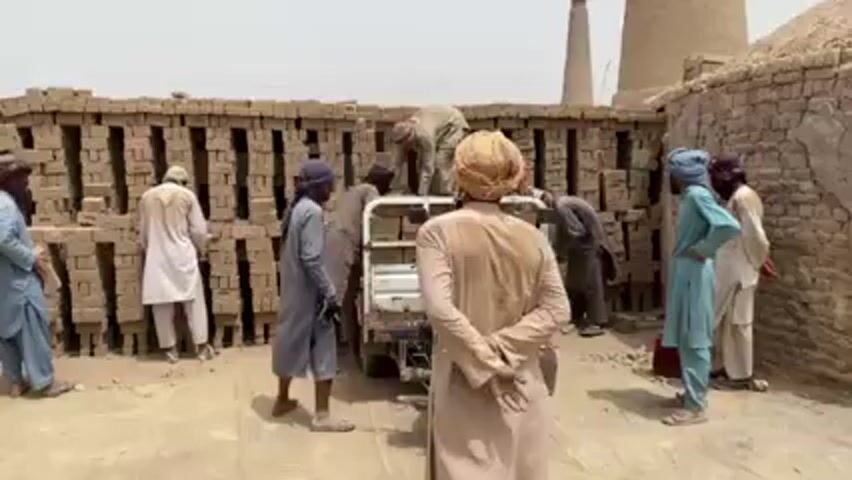 Several men stand near stacks of bricks in a dusty, outdoor setting. Some men are loading bricks from the stacks onto a small vehicle that appears to be a modified motorcycle with a flatbed. The men are dressed in traditional clothing, with turbans or head coverings. In the background, there are brick kilns. The day appears bright and sunny. Some men are talking as they work. One man stands with his back to the camera, observing the scene.