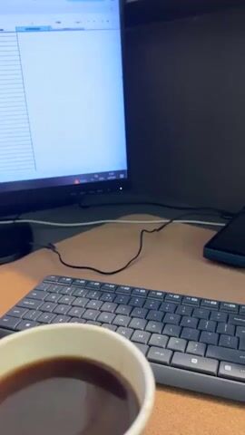 This is a close-up video of a desk with a computer monitor, keyboard, and a cup of coffee. The monitor displays a spreadsheet or table with blue and white columns. A hand holding a cardboard cup of coffee enters the frame from the bottom left and brings the cup closer to the camera. The coffee is dark brown. The background is blurred but appears to be an office environment with a dark partition behind the monitor. The video is short and has a slightly shaky camera movement.