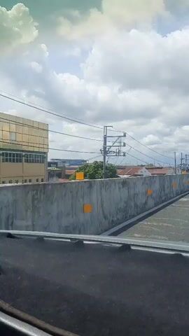 The video shows a view from inside a vehicle driving on an elevated roadway. The roadway has a concrete barrier on the left side, with orange markers at intervals. Beyond the barrier, there are buildings and trees. Overhead, there are power lines and utility poles. The sky is cloudy with patches of blue. In the distance, a speed limit sign of 40 is visible. The vehicle's dashboard is partially visible in the foreground.