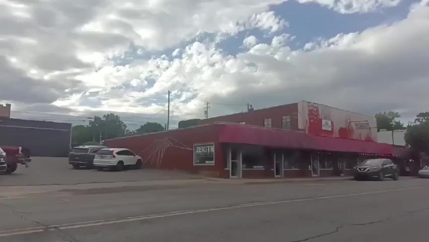The video shows a gray Toyota Tundra truck passing in front of a red brick building with a pink awning. The building appears to be a store or business. There are other cars parked in front of the building, and the sky is partly cloudy.