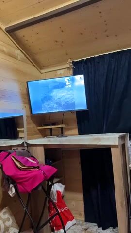 A television displays a weather report. On the screen, a man in a blue shirt and glasses points to a laptop displaying a map. Text on the screen identifies the man as Aleksei Pulin, a meteorologist. Behind the television, a wooden wall and a black curtain are visible. A black shelf with a green electronic device is mounted below the television. A small mirror is visible on the left side of the screen. A pink blanket and a white plastic bag are on a surface in the foreground.