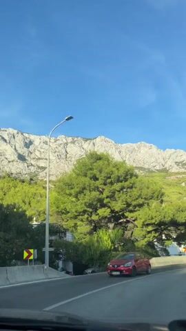 The video shows a view from inside a car traveling on a road. The road is paved and curves to the right. There are other cars on the road. On the left side of the road, there are trees and a large rock formation. The sky is blue with some clouds. The sun is shining. The video is taken from a first-person perspective, as if the viewer is driving the car.