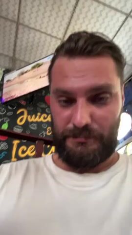The media file is a close-up, selfie-style video of a man with a beard and dark hair, wearing a white t-shirt. He is looking directly at the camera with various expressions.

In the background, a black sign with yellow lettering is visible, advertising "Juicy & Ice Bar." Various cartoonish icons of food items, such as burgers, apples, and ice cream cones, are also depicted on the sign. Above the sign, a television screen shows an image of a lion-like animal walking. The overall setting appears to be an outdoor or semi-outdoor establishment, possibly a cafe or bar, with a tiled ceiling and a fan visible on the right side of the frame.

The man's emotions shift throughout the short clip, from a neutral or slightly bored expression to one of surprise or amusement as his eyes widen. The lighting suggests it could be daytime, but the exact time of day is not definitively clear. The location context provided indicates Walasmulla, Sri Lanka. No specific activity or event is clearly discernible beyond the man interacting with the camera.