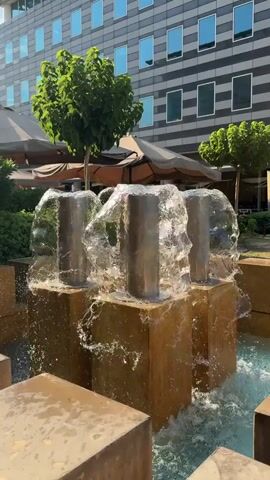 A fountain with three silver cylindrical spouts emerges from a blocky, golden structure, spraying water into a blue pool. In the background, a modern building with many windows and some trees and umbrellas are visible. The video is a short, close-up shot of the fountain.
