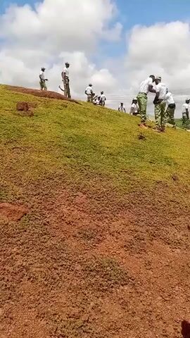 The video shows an outdoor scene with numerous young adults gathered on and around a grassy hill under a partly cloudy sky. The ground in the foreground and a path leading down the hill is reddish-brown dirt.

The subjects are dressed in a uniform consisting of white t-shirts, often with green trim, and khaki/green trousers. Many also wear green caps. In the foreground, a young man walks slowly from right to left, looking to his right. He is clearly visible, wearing a white t-shirt with "NYSC" printed in green on the chest, green trim on the collar and sleeves, khaki trousers, a green and white striped cap, and a green lanyard around his neck.

In the middle ground, on the grassy slope, several individuals are standing, sitting, and interacting. Beyond the slope, in the distance, a larger crowd of people is gathered on a flat, open area, some appearing to be in formations. Power lines are visible in the background, along with some distant buildings.

The scene appears to be a gathering or event, possibly a training exercise or assembly, involving members of the National Youth Service Corps (NYSC), as indicated by the uniform and visible text. The time of day is clearly daytime, with bright natural light and visible shadows. The setting is consistent with an open field or camp area in Durayabo, Nigeria.